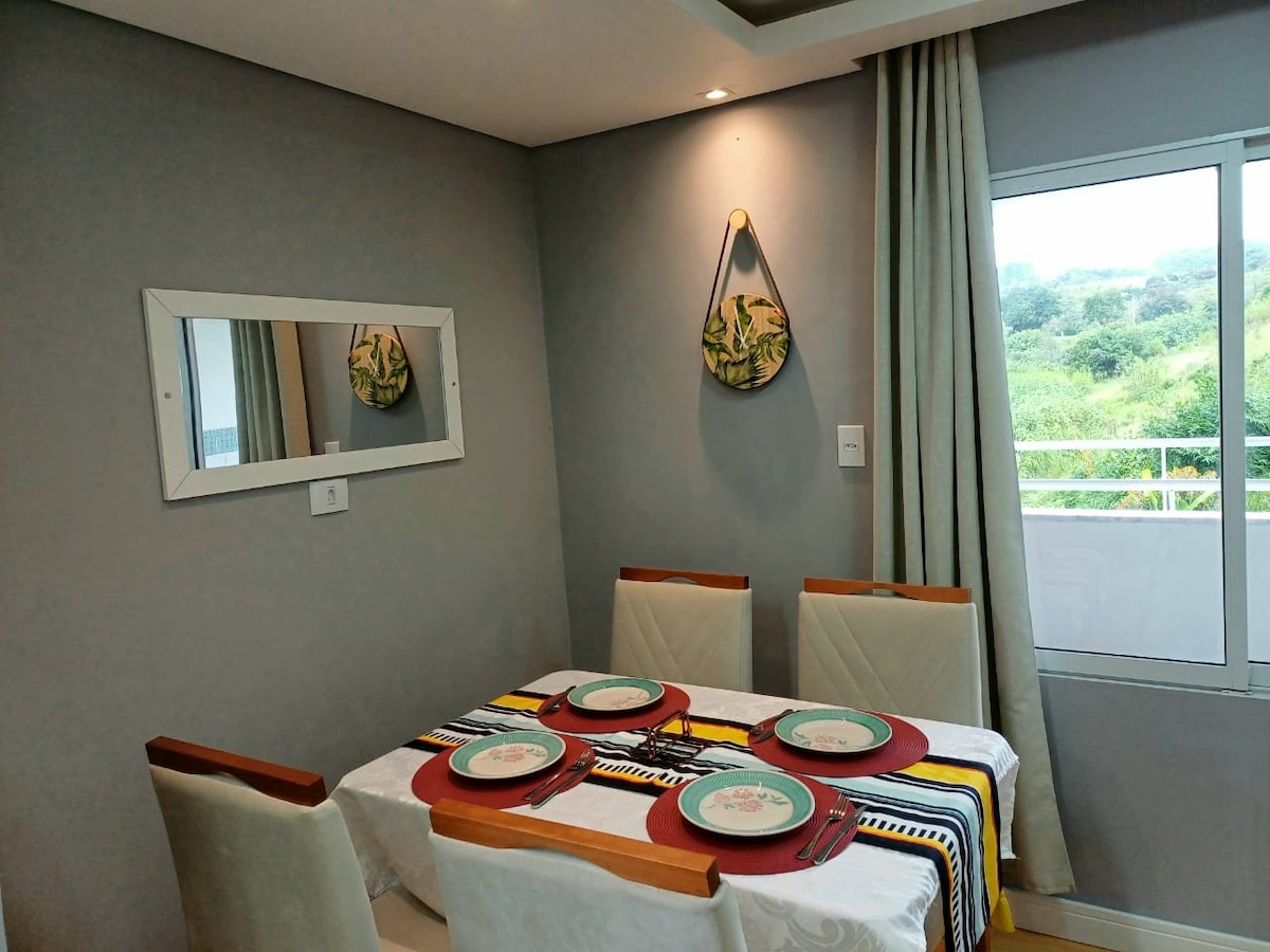 A dining area features a rectangular table set for four with colorful placemats. Light gray walls are adorned with a circular decorative piece and a mirror. Natural light enters through a large window, offering a view of the surrounding greenery.