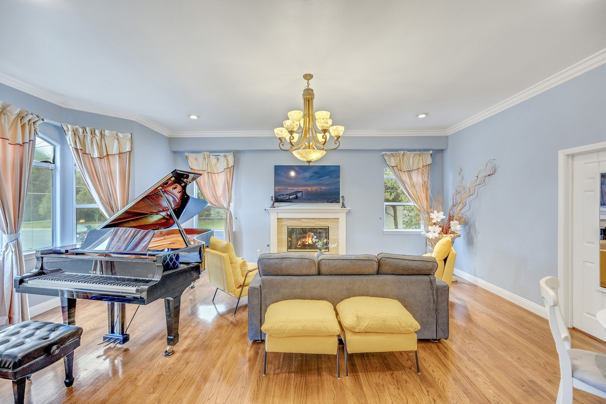 A spacious living area features a grand piano to the left and a cozy sofa facing a fireplace. Soft shades of blue adorn the walls, complemented by elegant curtains framing the windows. A stylish chandelier adds a touch of sophistication to the room.