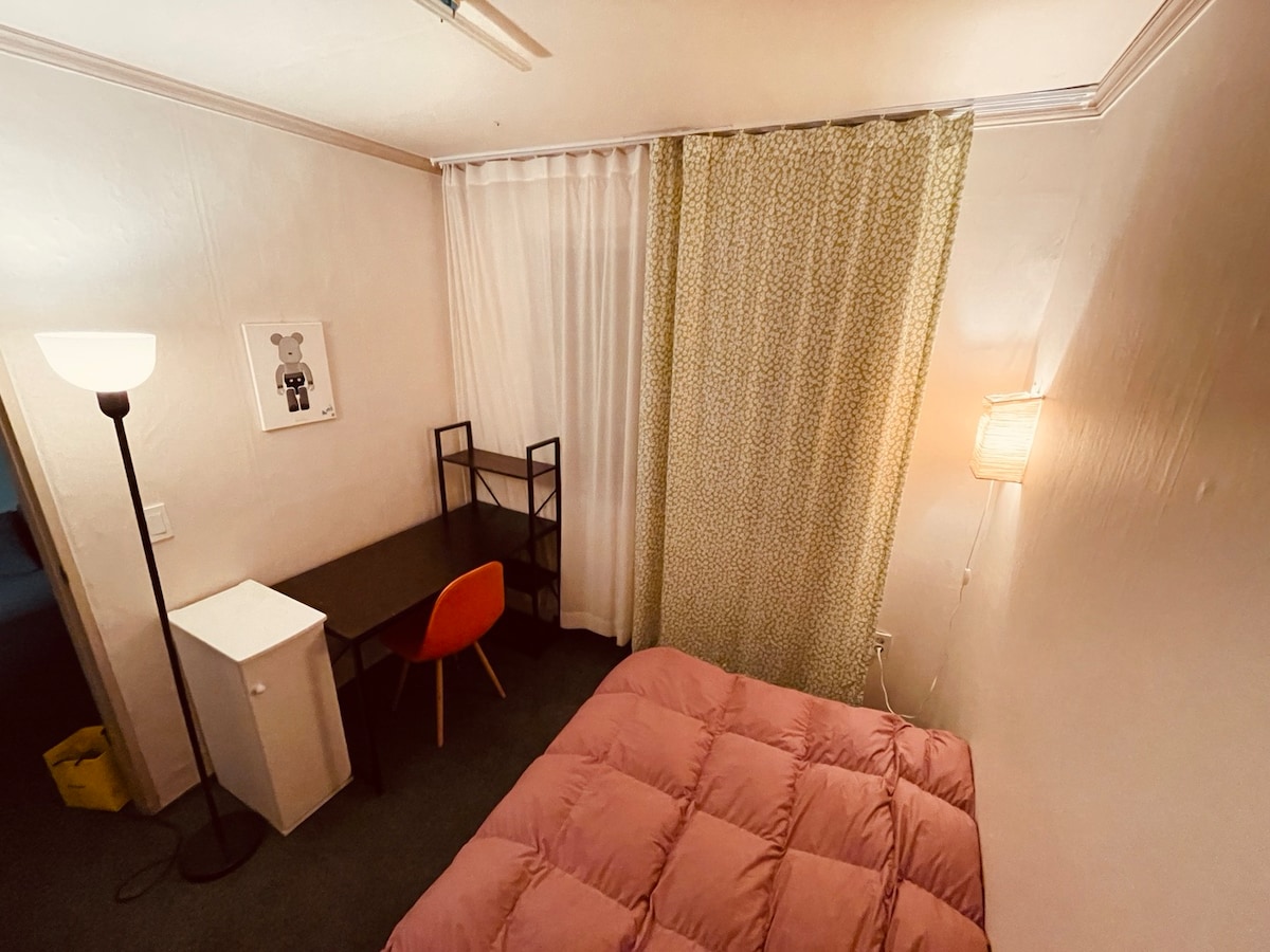 A simple room features a super single bed with a cozy pink comforter. A desk and chair are positioned adjacent to a window dressed with sheer and patterned curtains. Soft lighting is provided by a standing lamp, contributing to a calm atmosphere.