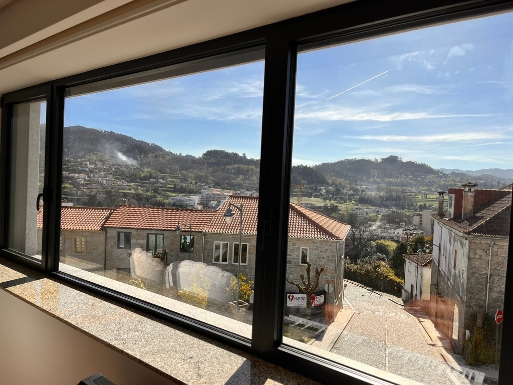 A large window provides a sweeping view of the surrounding landscape, featuring rolling hills and traditional stone houses. Bright sunlight fills the room, highlighting the modern window frame and the contrasting textures of the exterior stone walls.