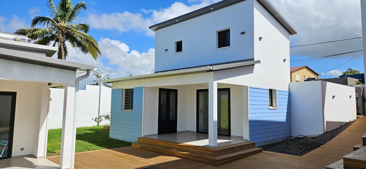 A two-story building features a modern design with blue and white accents. Large glass doors provide access to the front porch, surrounded by a wooden deck. Lush greenery is visible in the yard, complemented by palm trees against a bright sky.