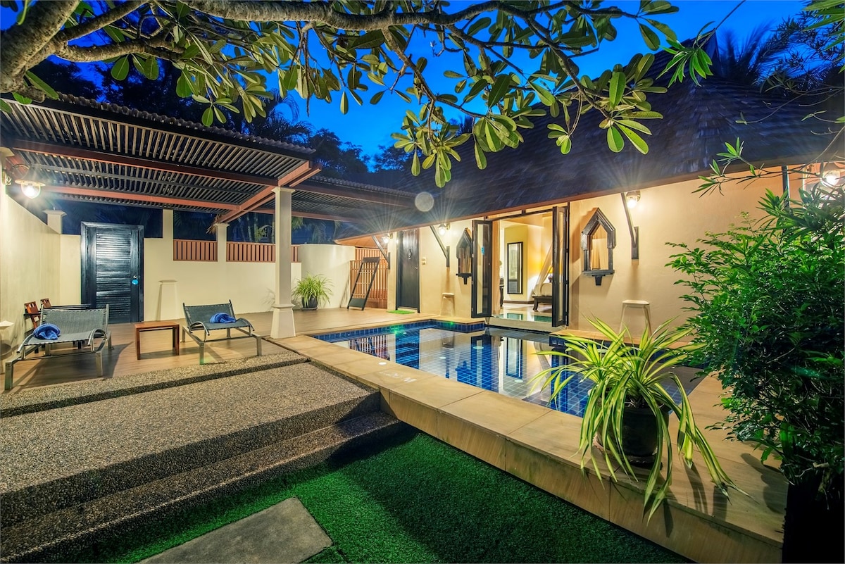 A serene private pool area is illuminated softly by outdoor lighting, featuring a blend of natural stone and lush greenery. Comfortable seating options are arranged beside the crystal-clear water, while elegantly designed villas are visible in the background under a clear evening sky.