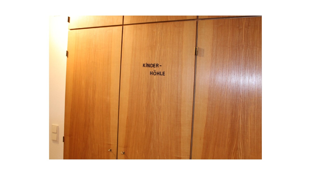 A wooden closet door is visible, featuring the word 'KINDERHÖHLE' prominently displayed. The grain of the wood adds texture to the surface, while the door handles are subtly integrated into the design.