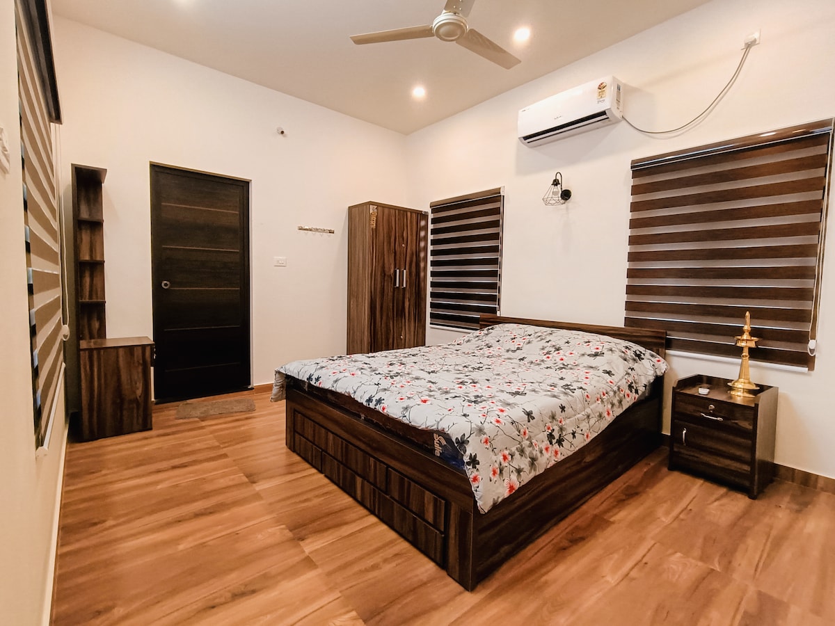 The room features a spacious bed adorned with a floral-patterned bedspread. Wooden floors enhance the natural light that enters through the two side windows, which are covered with dark blinds. A wardrobe and bedside table in matching wood complete the minimalist decor.