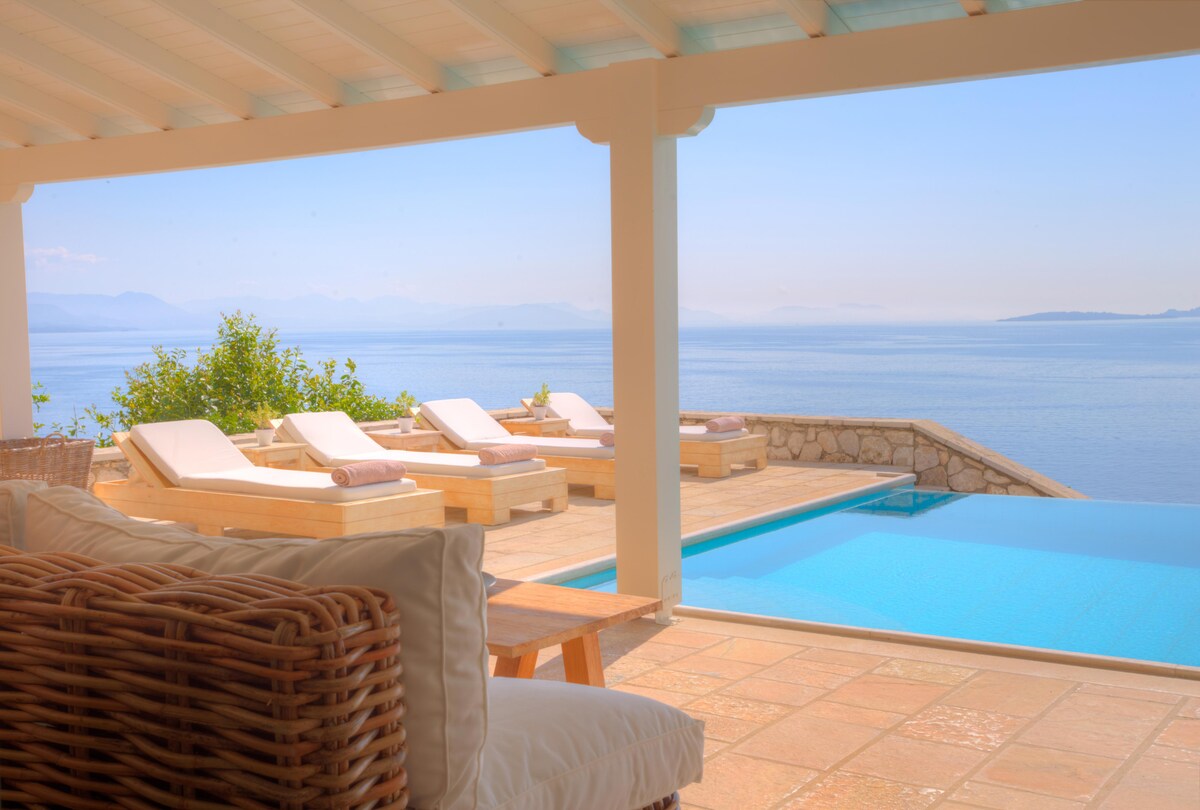 An outdoor terrace features a spacious seating area with woven furniture, overlooking a serene pool and ocean views. Sun loungers are arranged along the pool's edge, providing comfortable spaces for relaxation under clear skies.