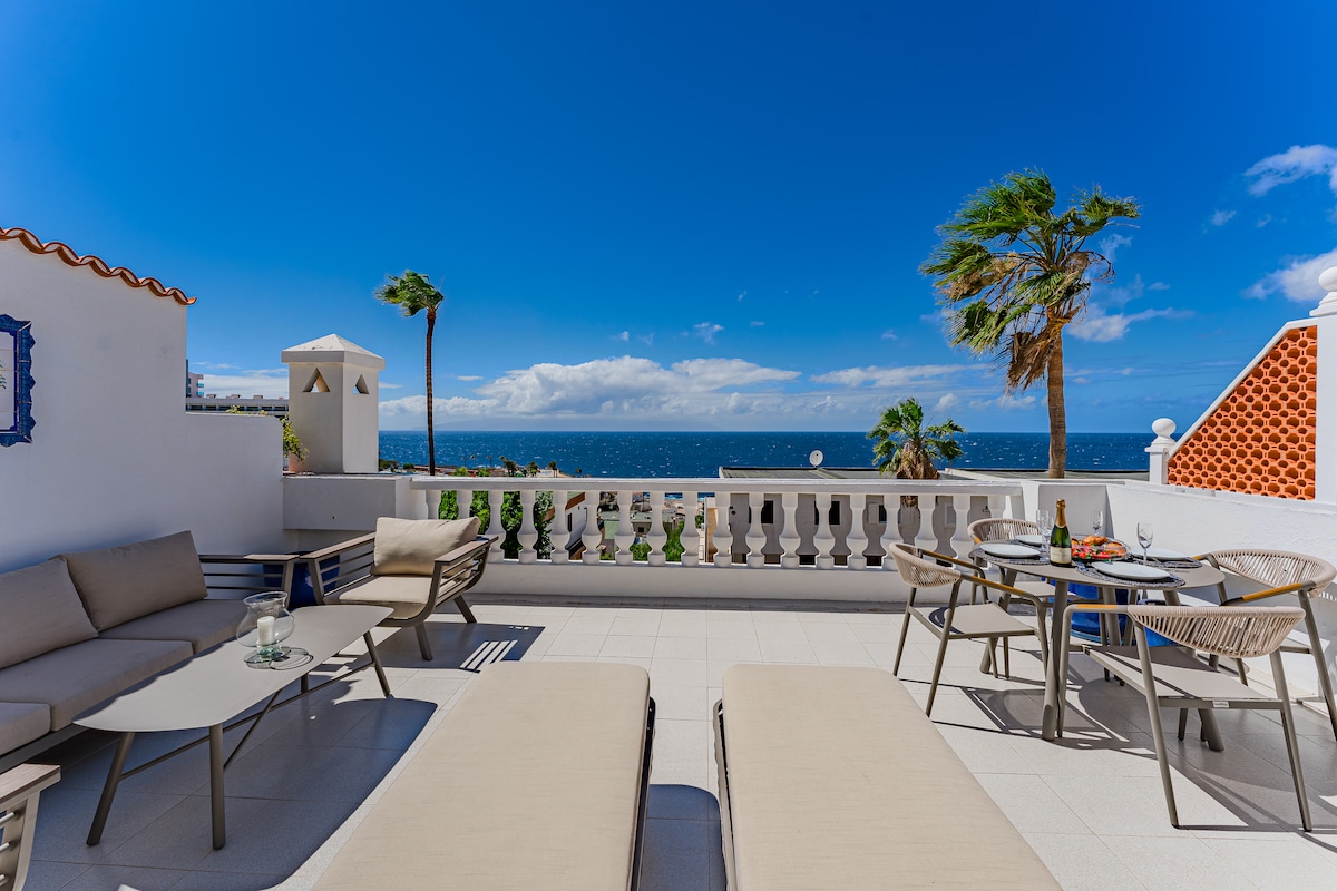 A spacious terrace features outdoor seating, a dining table, and two large sun beds set against a backdrop of ocean views. Lush palm trees sway gently in the breeze under a clear blue sky, providing a serene setting for relaxation and dining.
