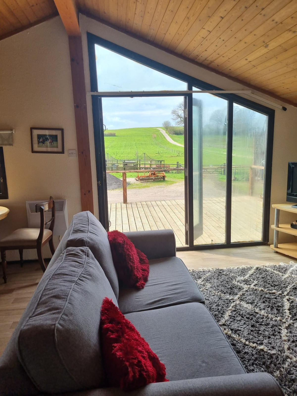 A spacious living area features a comfortable grey sofa with red cushions, positioned near large glass doors that open to scenic views of rolling green hills. The wood-paneled ceiling adds warmth, while a patterned rug enhances the cozy environment.