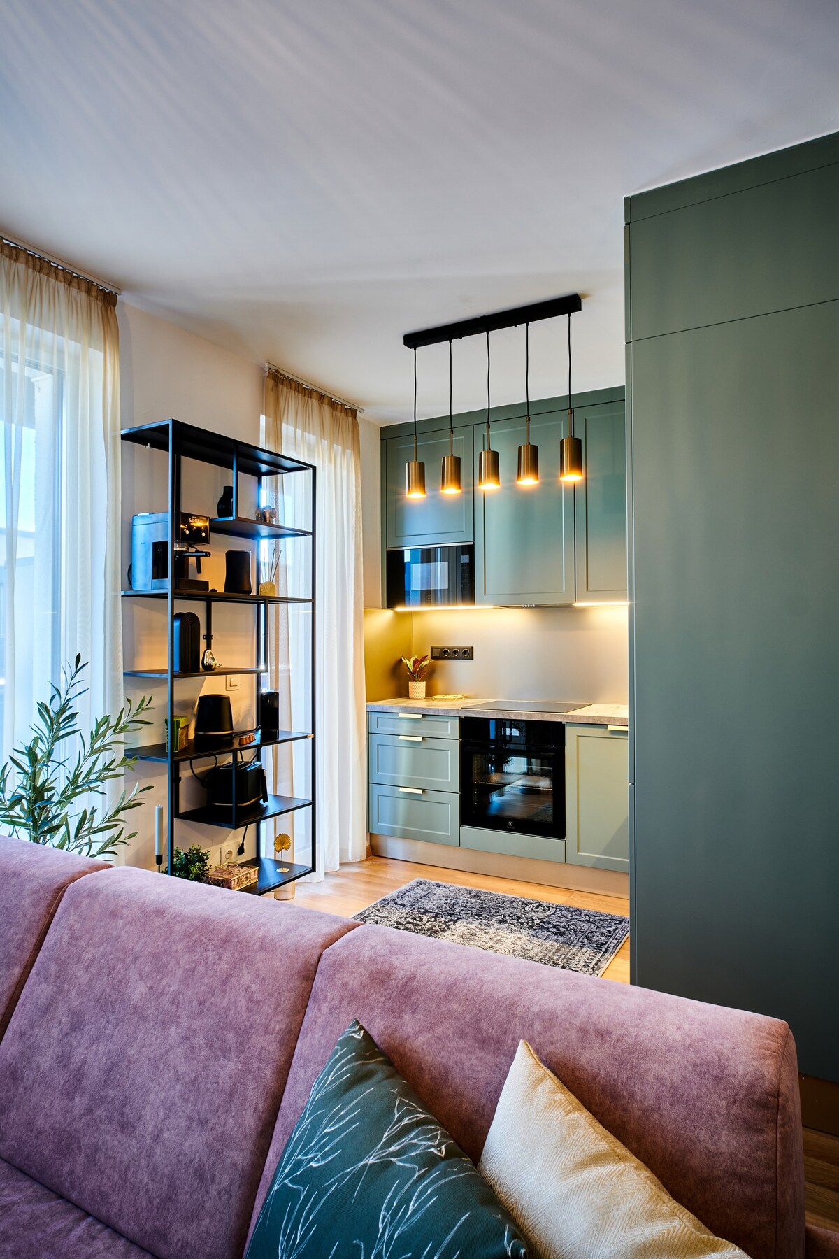 A modern kitchen is presented, featuring soft green cabinets and warm lighting. An open shelf holds various kitchen appliances and décor, while a cozy seating area with a pink sofa and decorative pillows is visible in the foreground. Large windows allow natural light to fill the space.