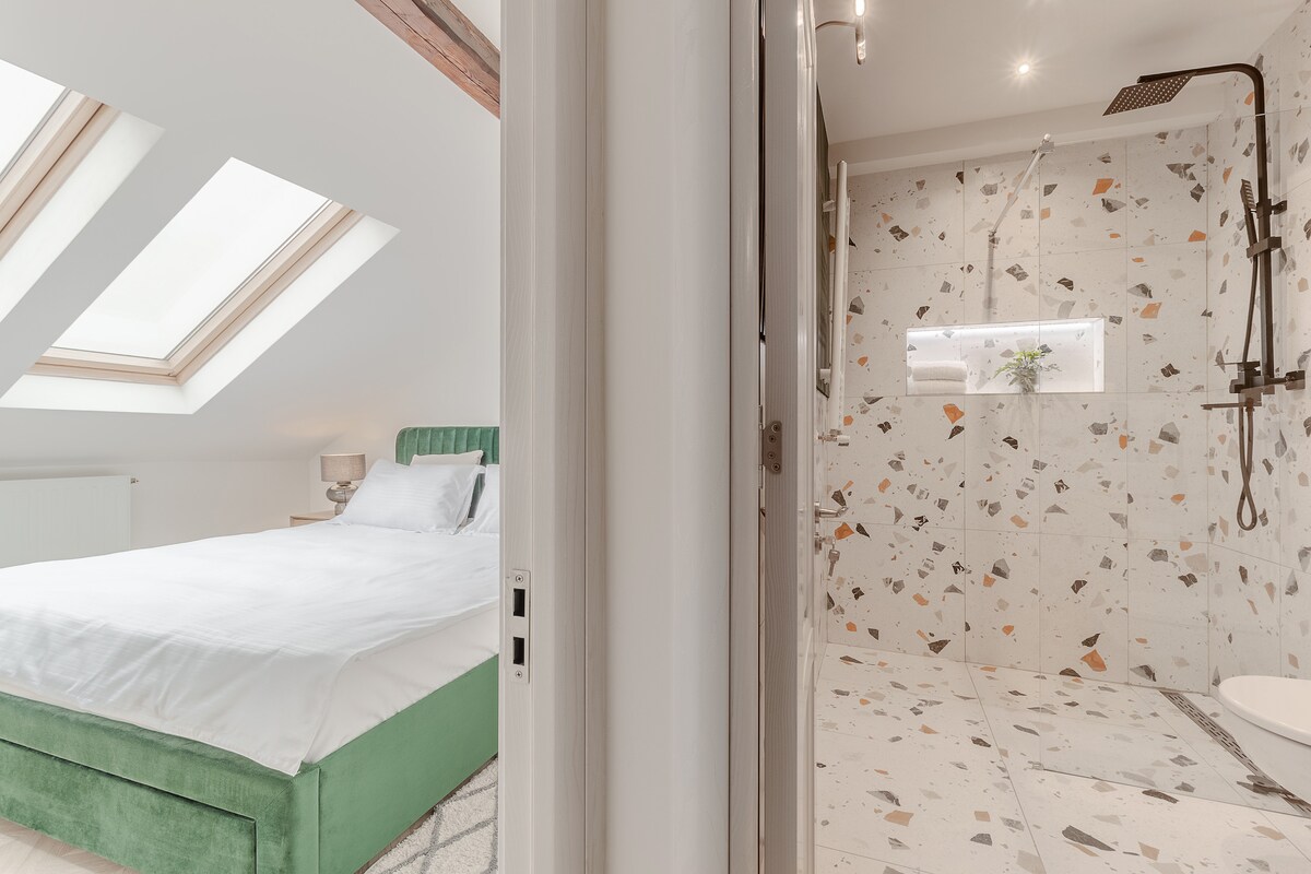 A bright bedroom features a spacious bed with a light-colored duvet and an elegant green headboard. Skylights allow natural light to fill the room. A modern bathroom is visible, showcasing a walk-in shower with a rain showerhead and a decorative tiled wall.