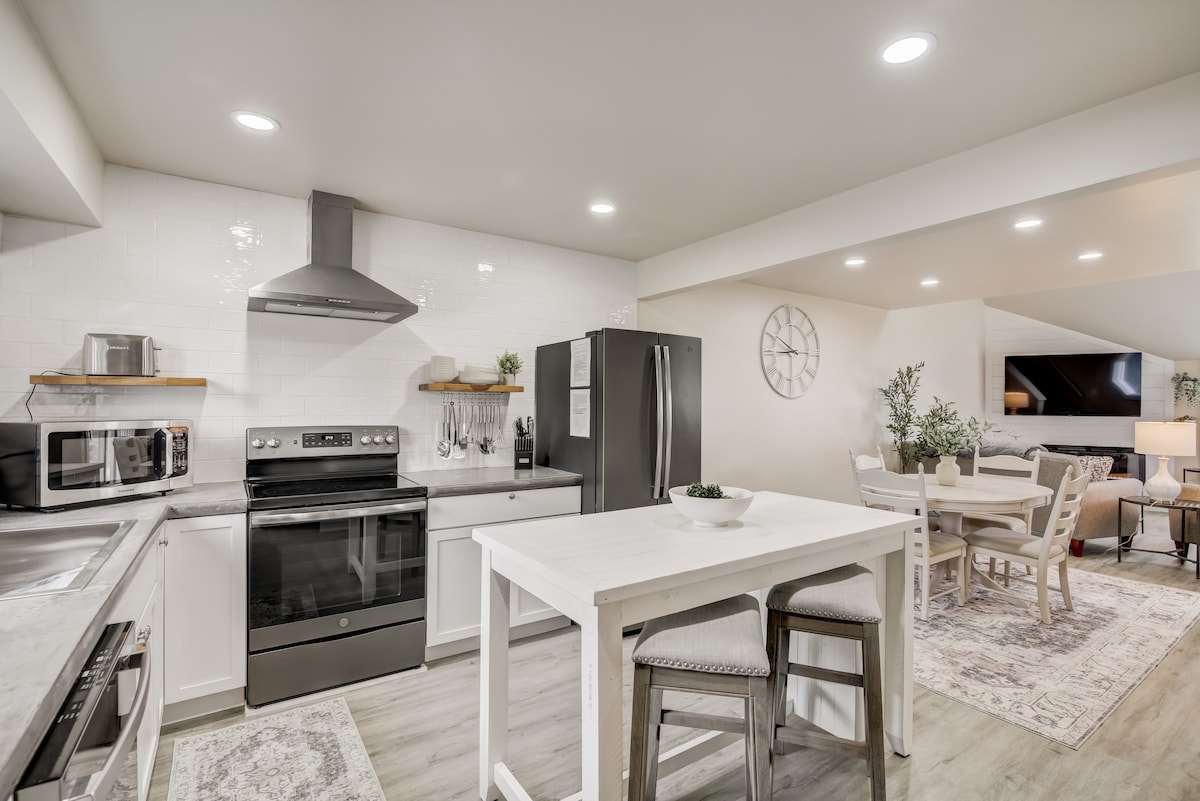 The kitchen features sleek white cabinetry and modern appliances, including a stainless steel oven and a microwave. A dining table is positioned nearby, surrounded by comfortable chairs. Natural light illuminates the space, complementing the neutral color palette.