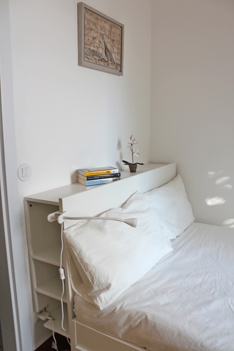 A cozy sleeping area is presented, featuring a spacious bed with white linens. A low headboard serves as a shelf, displaying books and a small decorative plant. A bedside lamp is attached for reading, while a picture frames a soft wall.