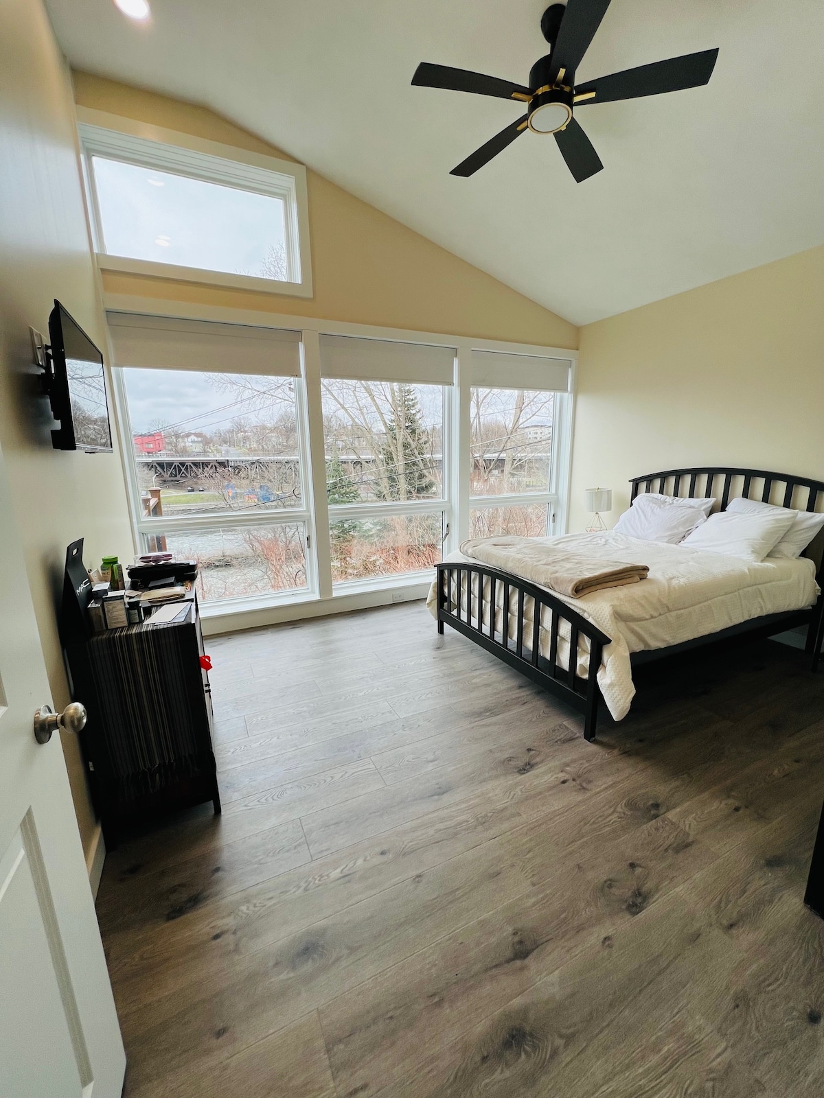 A bright bedroom features large windows allowing natural light to fill the space. The room is furnished with a dark wooden bed dressed in white linens, alongside a small black nightstand. A ceiling fan is positioned above, enhancing airflow, and a stylish dresser is located near the door.