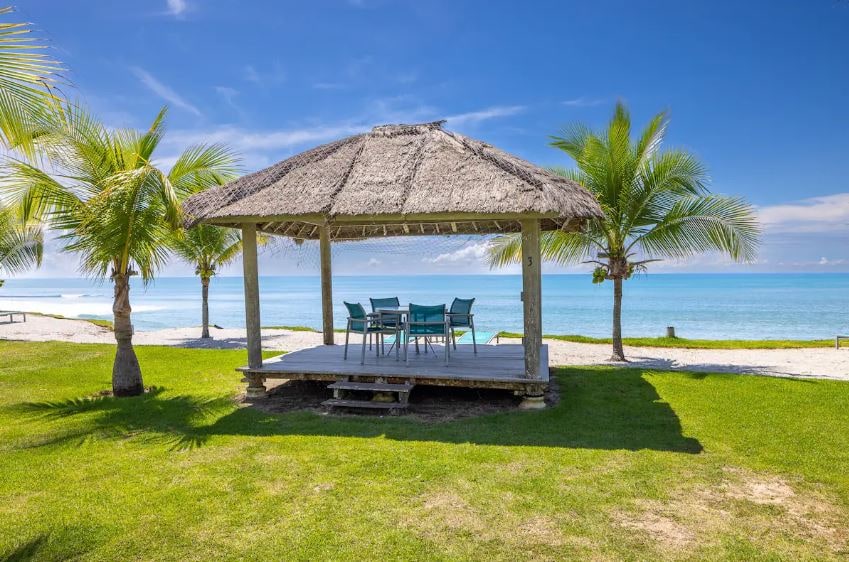 A thatched-roof pavilion is positioned on a grassy area overlooking the beach. A large dining table surrounded by six chairs invites outdoor meals. Lush palm trees frame the scene, with the ocean visible in the background under a clear blue sky.
