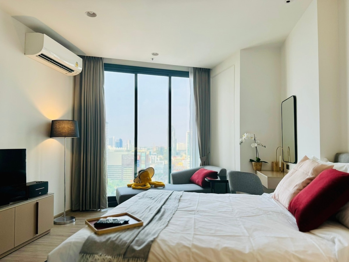 A modern guest room features a spacious bed with gray and red accents. A large window offers a view of downtown Pattaya. A television sits across from the bed, and a comfortable seating area with a gray armchair complements the relaxing atmosphere.
