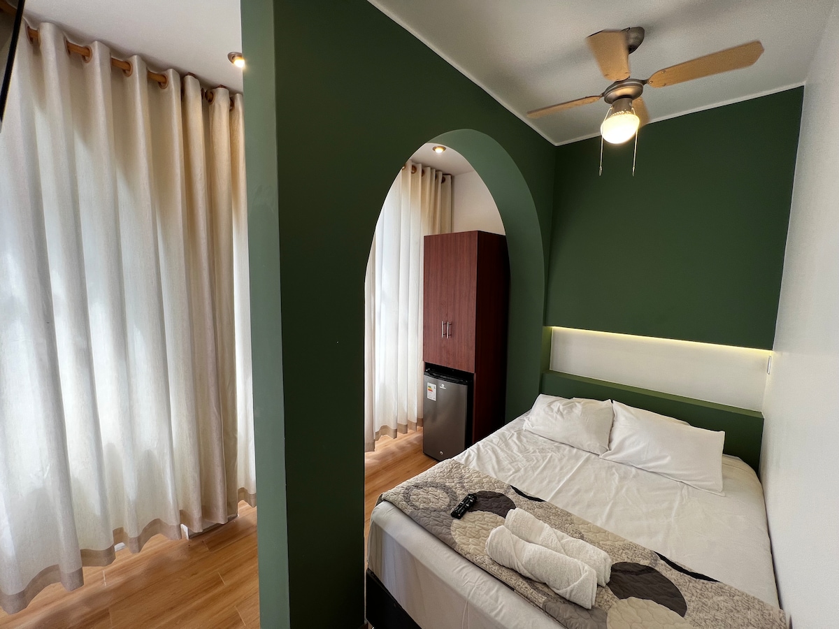 A cozy bedroom features a spacious bed with white linens, complemented by soft towels. An archway leads to a bright area with large windows adorned with light curtains. A brown cabinet and a small refrigerator are visible, enhancing the room's functionality.