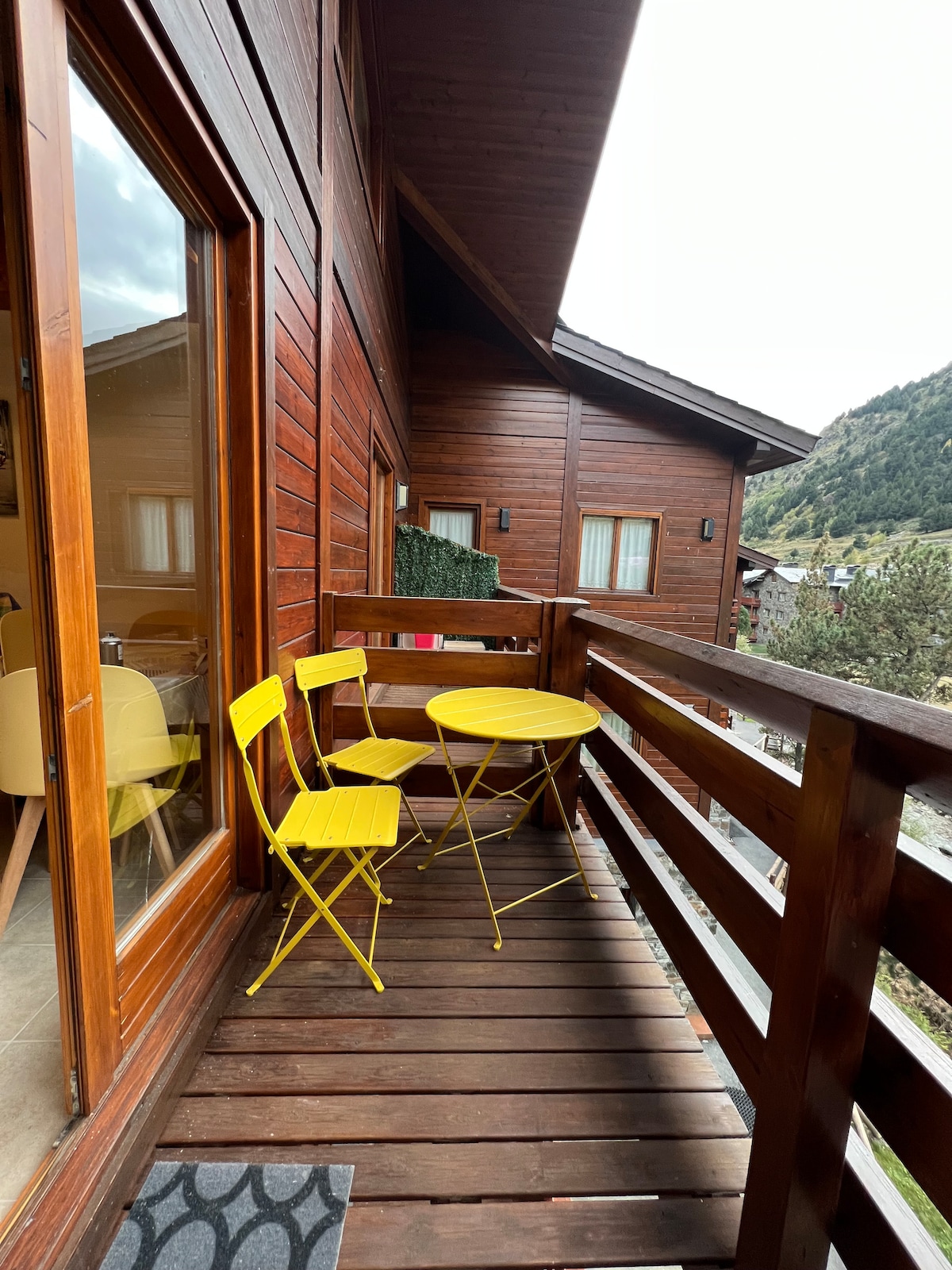A small balcony features yellow bistro-style furniture, consisting of a round table and two chairs. The wooden railing and walls provide a warm, rustic appearance, while views of the surrounding natural landscape are visible through the glass doors.