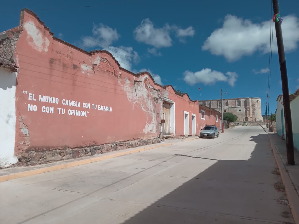 A quiet street is lined with colorful colonial buildings. A mural with text is visible on one wall, and a parked car sits along the curb. The sky is bright blue, scattered with clouds, enhancing the charming atmosphere of the town.