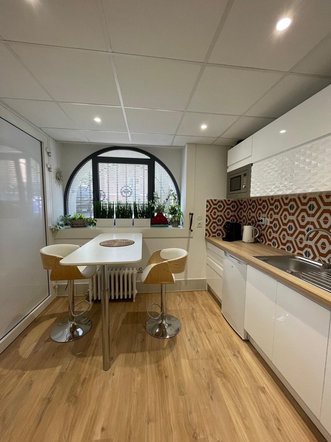 A modern kitchen features a sleek white cabinetry design and a distinctive patterned backsplash. A round table accompanied by two white bar stools is positioned near a large arched window, allowing natural light to illuminate the space. Additional appliances include a microwave and a refrigerator.
