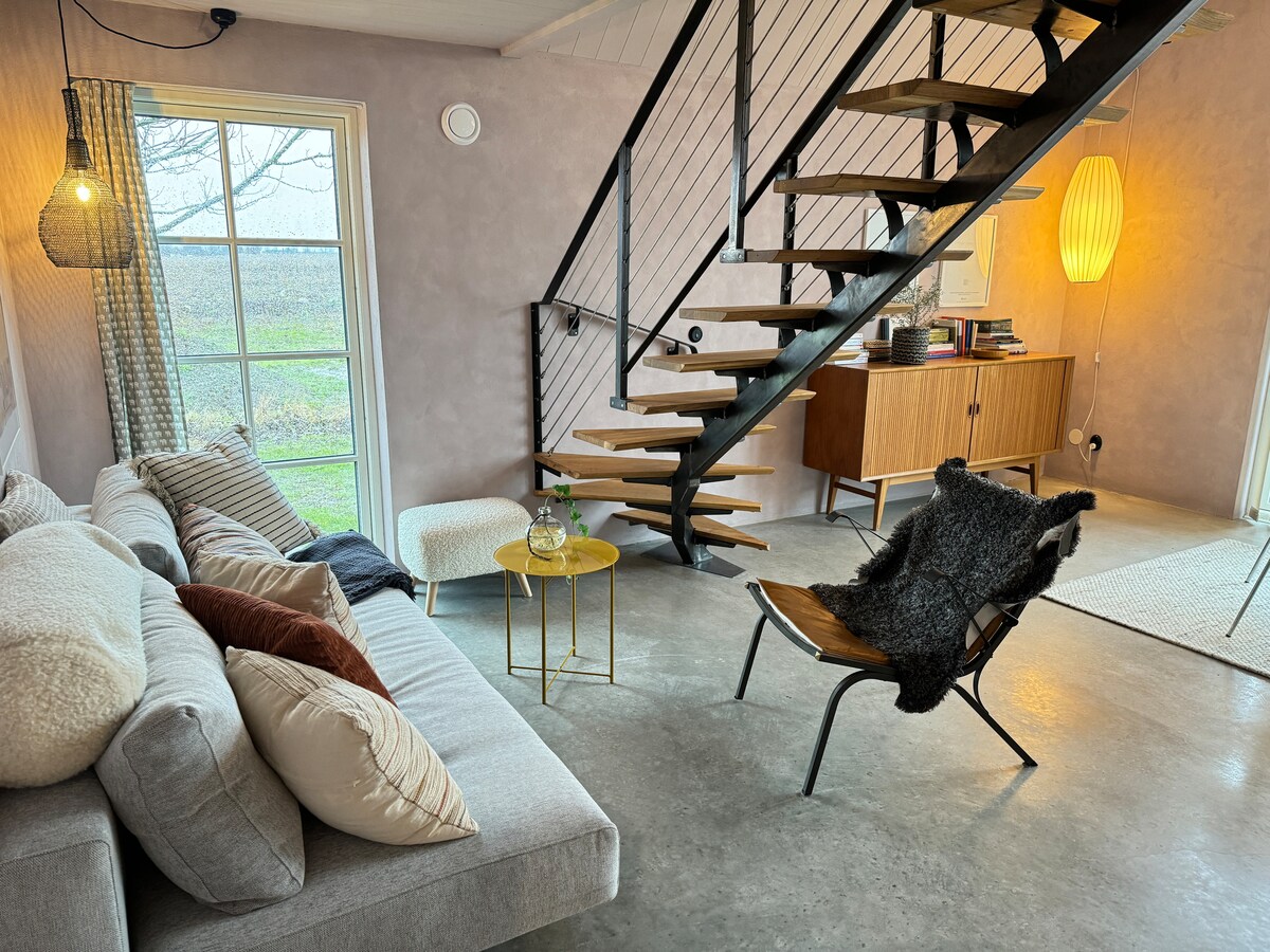 A cozy living area features a light grey sofa accompanied by accent pillows in warm tones. A sleek staircase with wooden steps leads to the upper levels. A small table holds refreshments, while a large window offers a view of the outdoor landscape.