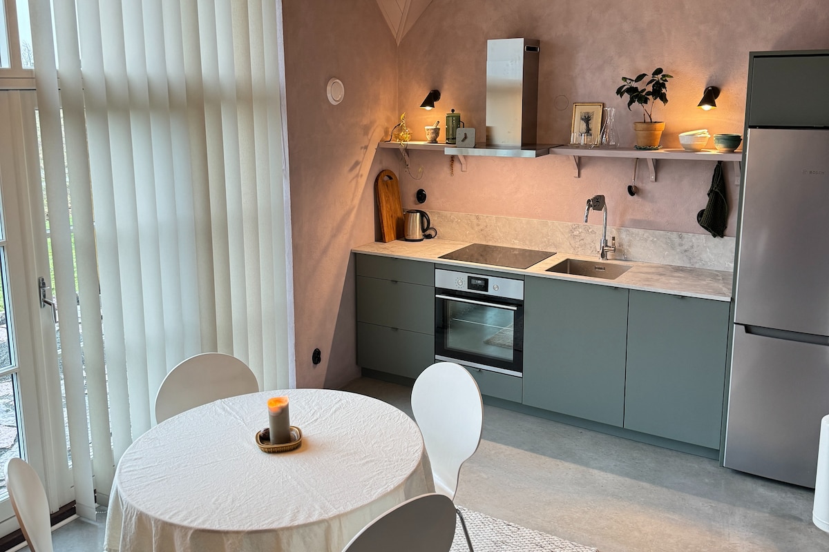 A modern kitchen space is equipped with sleek, green cabinetry and a marble countertop. A round table with white chairs is positioned nearby, complemented by soft lighting. Large windows with vertical blinds allow natural light to fill the area, enhancing its spacious feel.