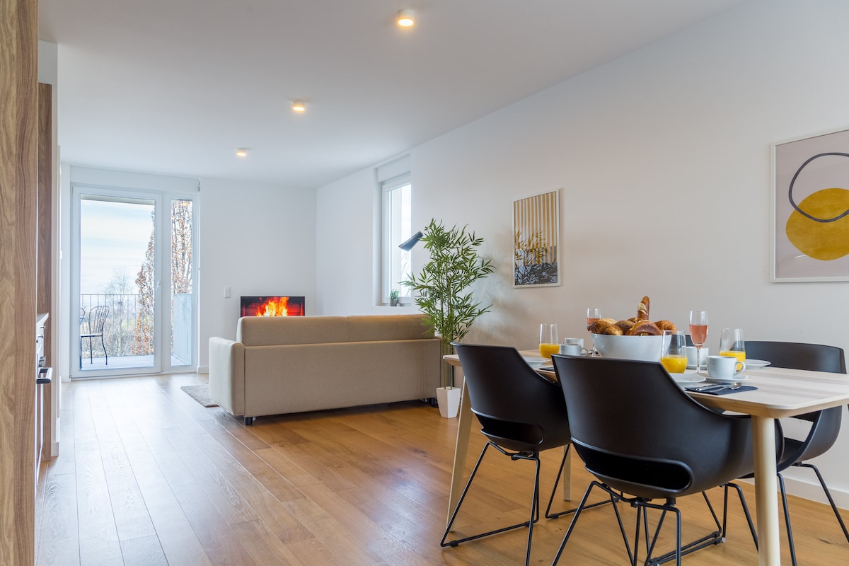 A bright and spacious living area features a comfortable sofa facing a cozy fireplace. A dining table is set with breakfast items, including drinks and pastries. Large windows allow natural light to fill the room, complementing the modern decor.