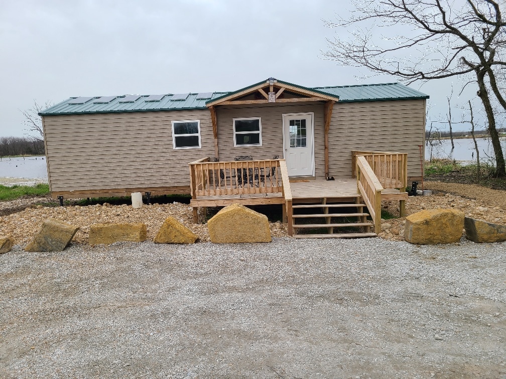 The exterior of a cabin is featured, showcasing a wooden deck with stairs leading up to the entrance. Surrounded by gravel, large boulders frame the area, while a lake is visible in the background along with overcast skies.