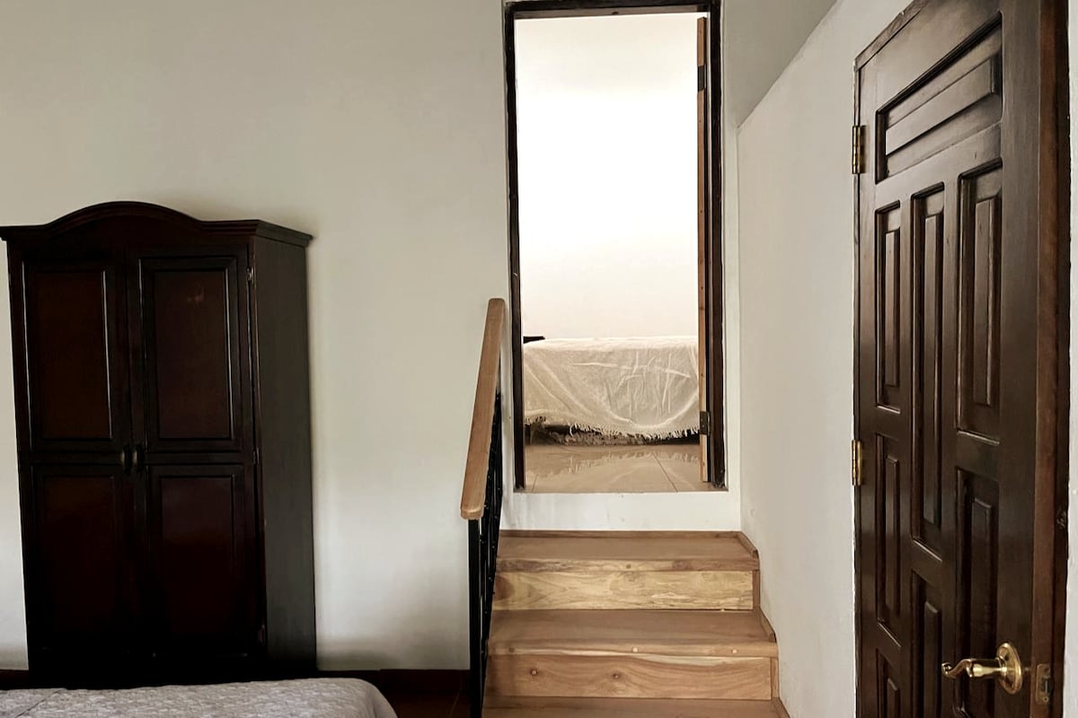A staircase leads to the upper level, visible beyond a wooden door. A dark wood wardrobe is positioned to the left, providing ample storage. The walls are painted in a neutral tone, and soft, natural light fills the space.