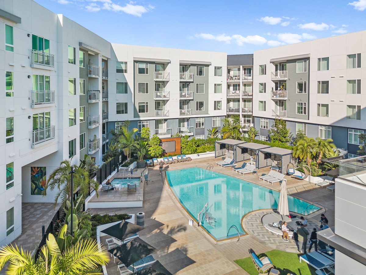 A spacious outdoor pool area is surrounded by modern apartment buildings. Sun loungers line the poolside, and cabanas provide shaded seating. Lush greenery and palm trees enhance the inviting atmosphere, while a hot tub is visible near the pool, promoting relaxation.