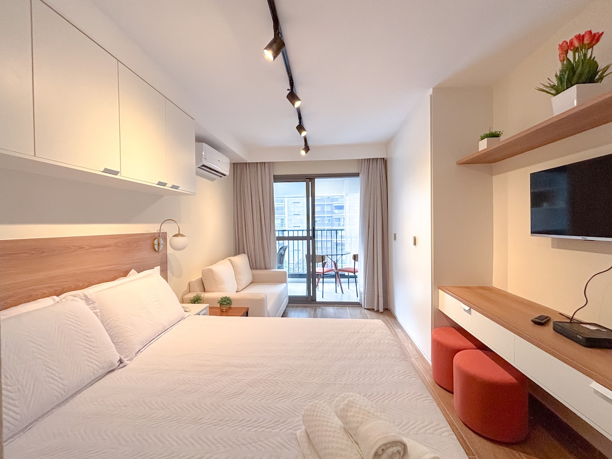 A studio layout is depicted with a spacious bed positioned against a light wood accent wall. A sofa and a small balcony with seating are visible, while a wall-mounted television and a decorative shelf complete the room's furnishings. Natural light enters through sliding glass doors.