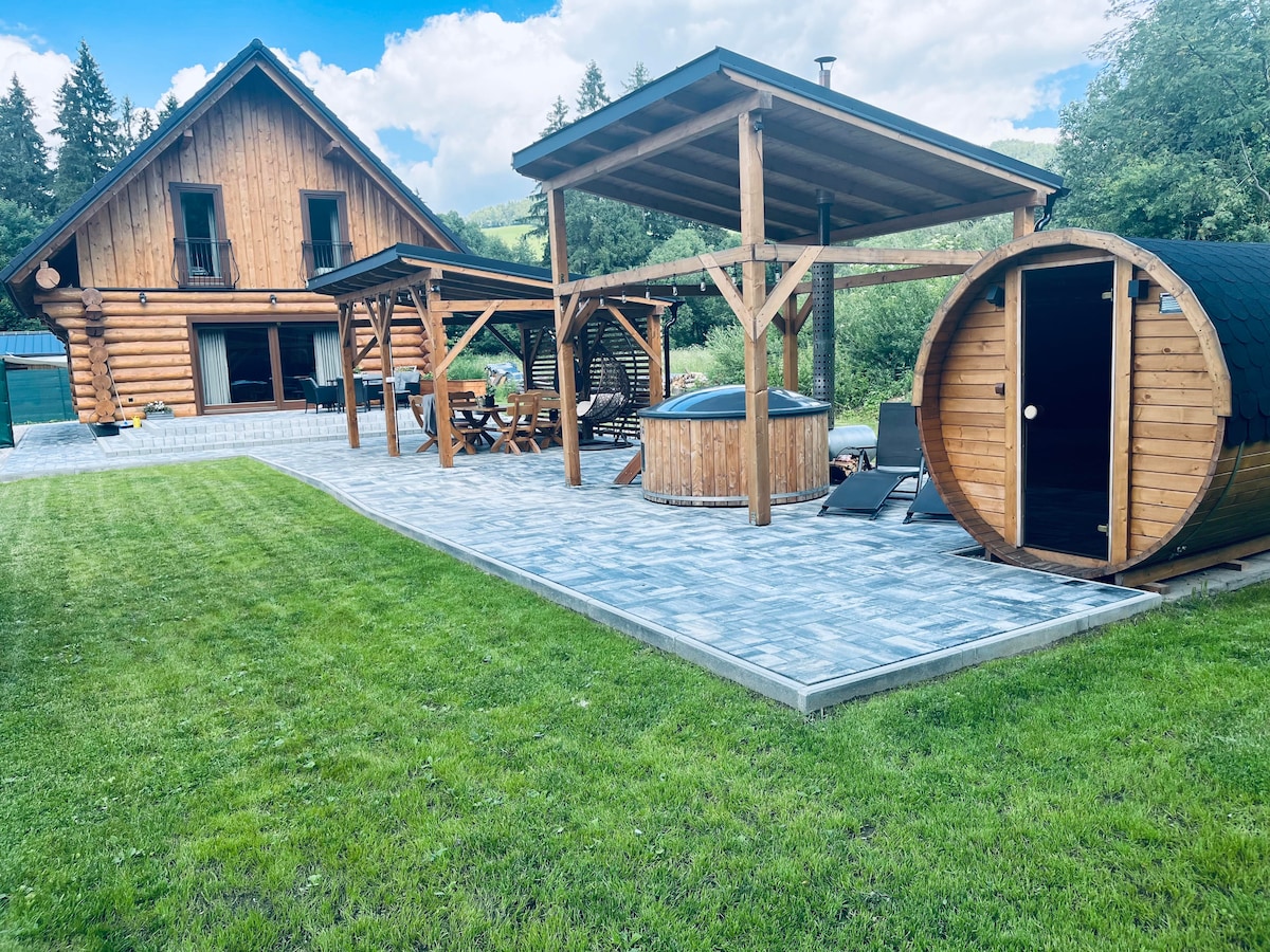 An outdoor area features a well-maintained lawn with a stone patio, showcasing a hot tub and a wooden sauna. Sheltered seating areas are provided under wooden structures, and the rustic log cabin is visible in the background, surrounded by trees.