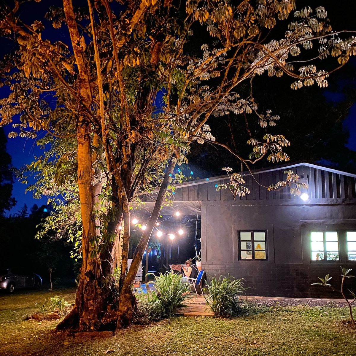 A cozy cabin is set against a backdrop of greenery, illuminated by soft outdoor lighting. A porch is visible, with seating and decorative plants. A large tree provides shade, while the evening sky enhances the tranquil atmosphere of the setting.