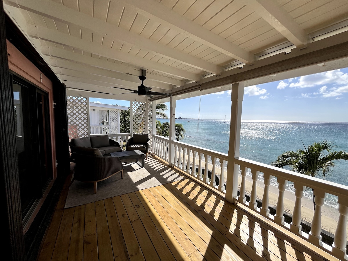 A covered patio area features a spacious seating arrangement with comfortable chairs and a coffee table, offering a view of the ocean and palm trees. Sunlight reflects off the water, creating a serene coastal atmosphere.