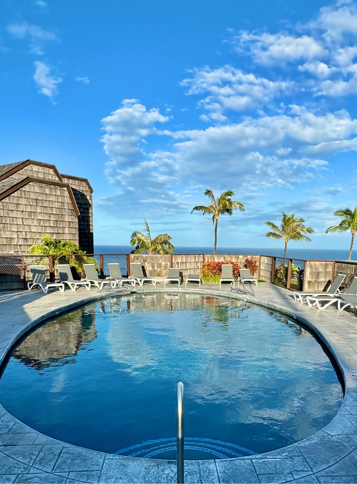 A circular pool with a tiled edge reflects the blue sky and fluffy white clouds. Surrounding the pool are lounge chairs positioned neatly along the perimeter, with tropical plants and palm trees visible in the background, adding a touch of greenery.