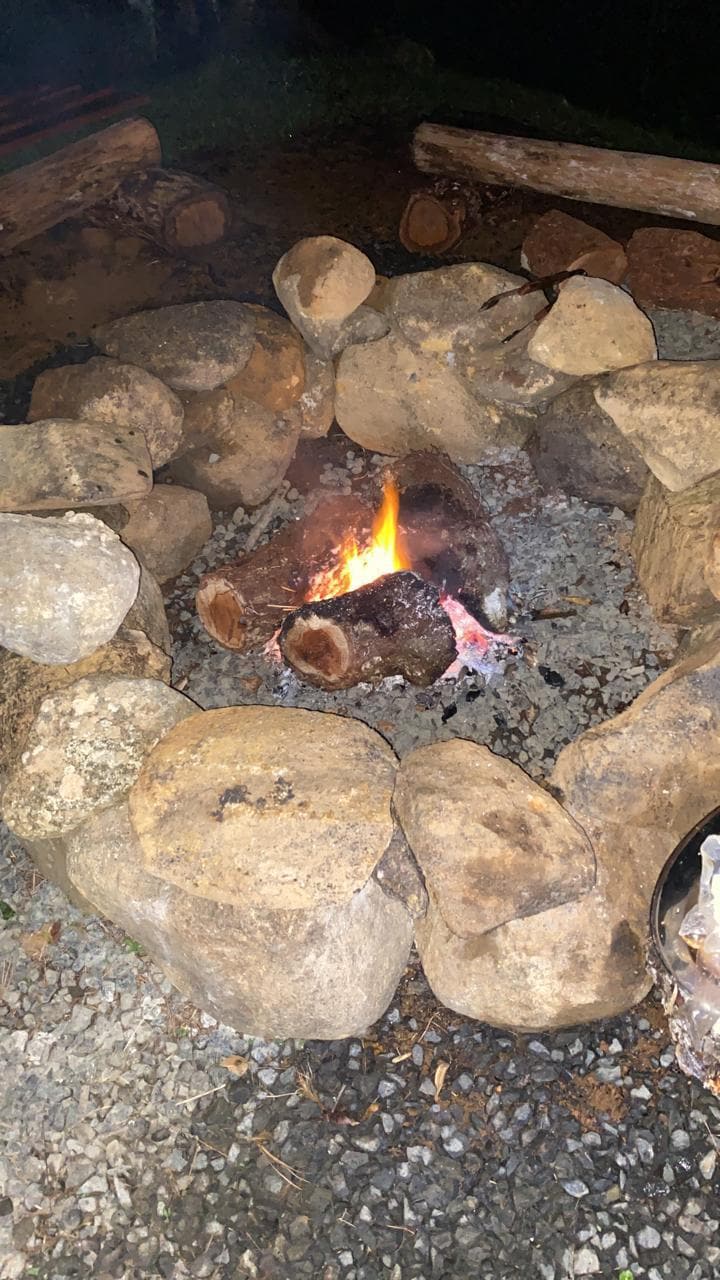 A fire pit encircled by irregular stones is seen, with flames flickering among burning logs. The surrounding gravel indicates a rustic setting, inviting gatherings in an outdoor environment. The glow of the fire contrasts softly with the darkness around.