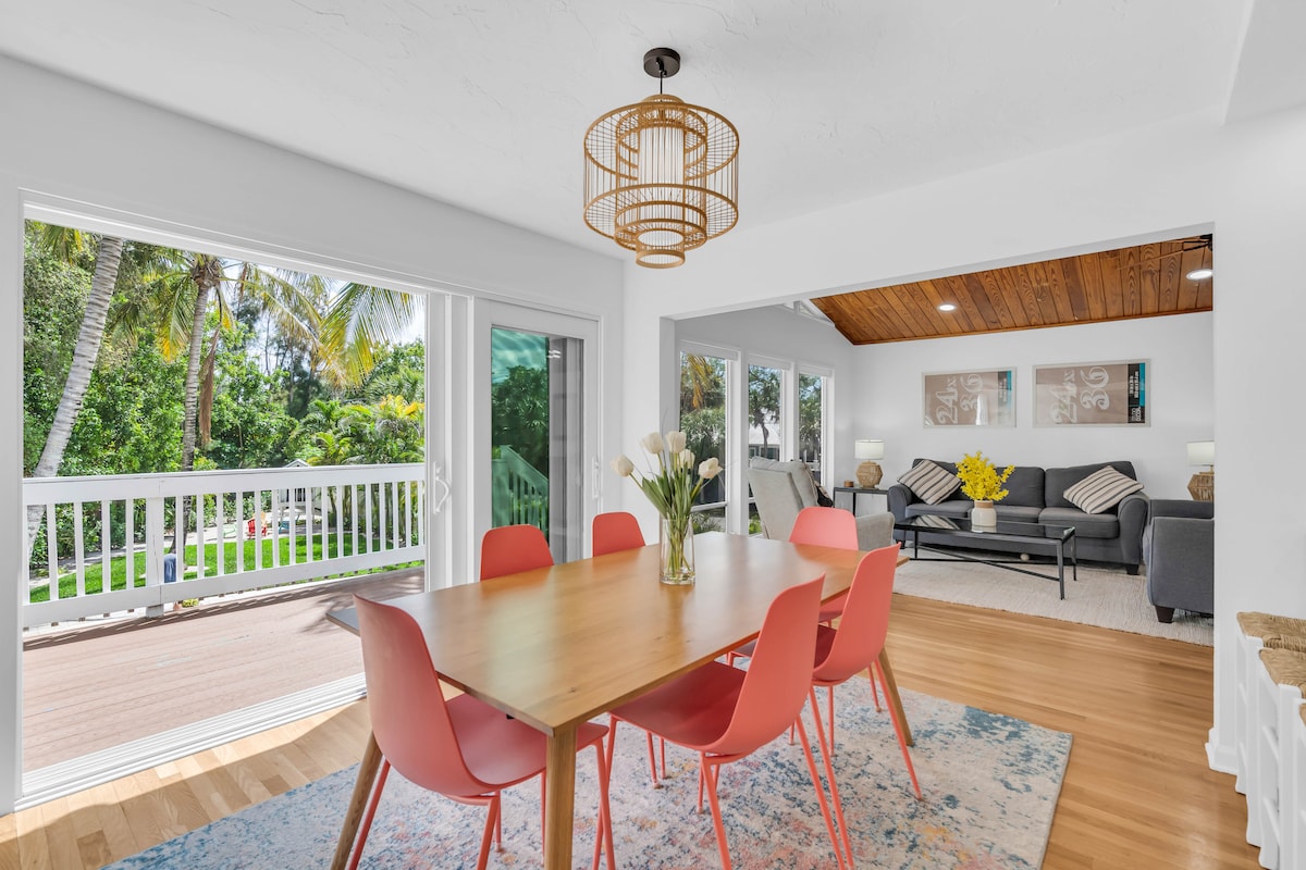 A bright dining area is highlighted by a wooden table surrounded by pink chairs, with a vase of tulips at the center. Large sliding doors provide views of the lush backyard, while natural light brightens the space, enhancing the inviting atmosphere.