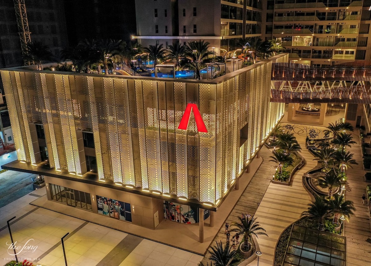 A modern building is highlighted by intricate façade patterns and soft lighting. Palm trees surround the entrance, which is paved with light-colored tiles. A swimming pool area is visible above, creating a welcoming atmosphere in the vibrant urban environment.