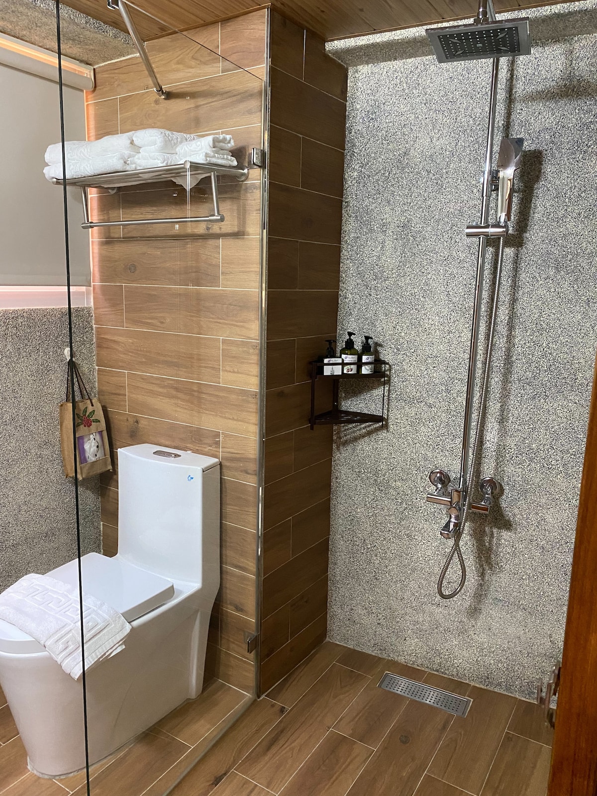 The bathroom features a modern design with wooden accents and stone-textured walls. A shower with a rain showerhead is present alongside a sleek toilet. Towels are neatly arranged on a hanging rack above, while a small shelf holds toiletries.
