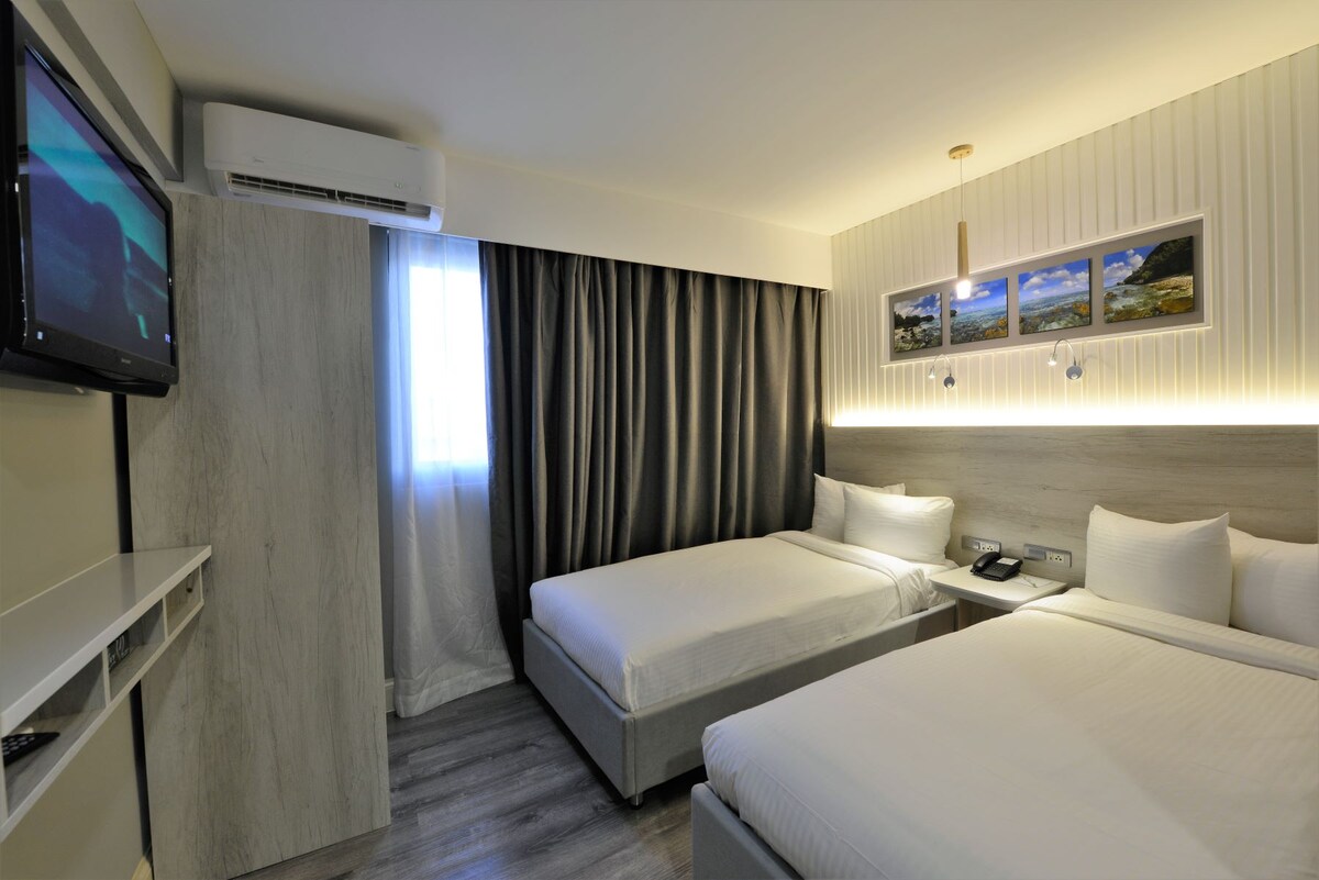A modern guest room features two spacious beds with white linens, a wall-mounted television, and a light wood accent wall. A window with sheer curtains allows natural light to fill the space, complemented by dark drapes for privacy. Decorative artwork is showcased above the beds.