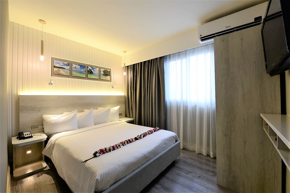 A studio room is configured with a spacious bed, elegantly decorated with flower arrangements. Soft lighting creates a calming ambiance, while a large window provides natural light, and curtains frame the view. An air conditioning unit is installed above for comfort.