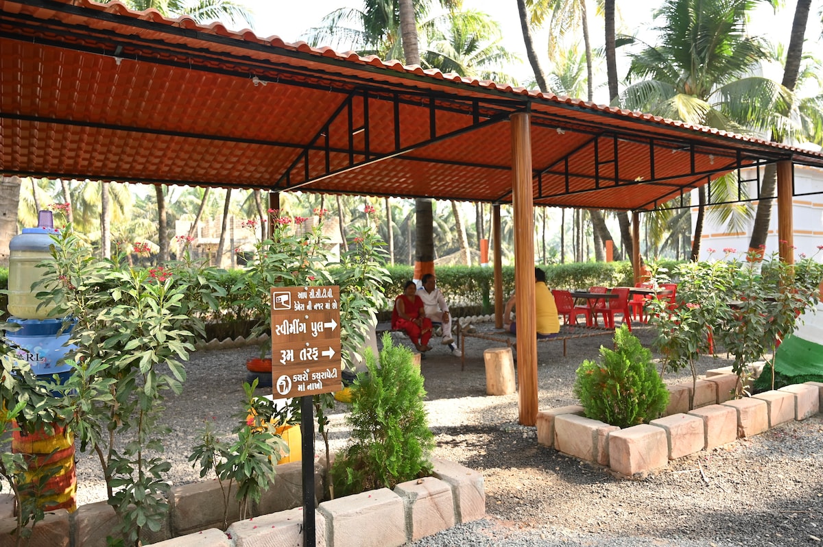 An open seating area is surrounded by lush greenery, featuring a rust-colored roof supported by black metal beams. Red chairs and wooden tables are positioned beneath the shelter, creating a relaxed space for guests to enjoy the outdoor setting.