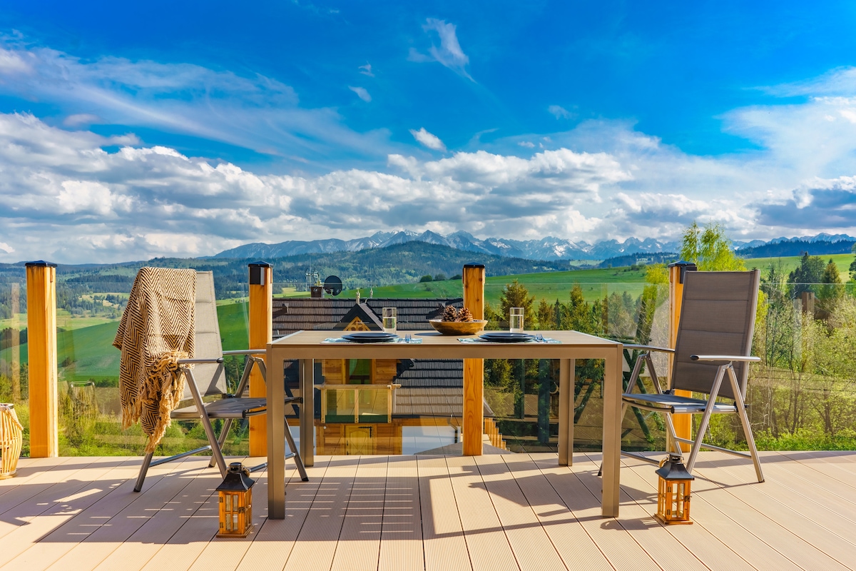 A spacious outdoor terrace features a modern table with two chairs, set against a backdrop of sweeping mountain views. Soft blankets are draped over one chair, while lanterns provide subtle lighting. The sky is bright with clouds, emphasizing the scenic landscape.