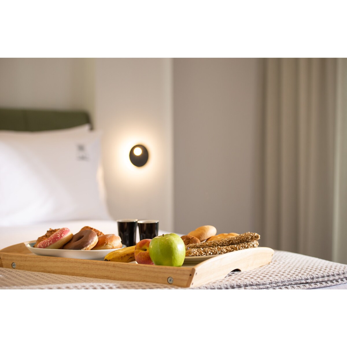 A wooden tray is positioned on a light-colored bedspread, displaying a variety of breakfast items, including pastries, an apple, and two cups. Soft lighting is provided by a wall-mounted fixture, complementing the neutral tones of the room.