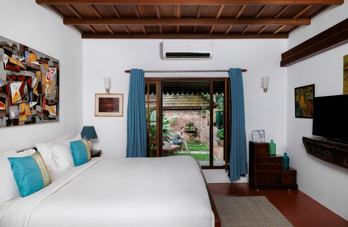 A bright bedroom features a spacious bed with white linens and colorful accent pillows. Large windows provide views of the outdoor garden area. Wooden beams and a warm palette create an inviting atmosphere, while art pieces adorn the walls, enhancing the visual interest.