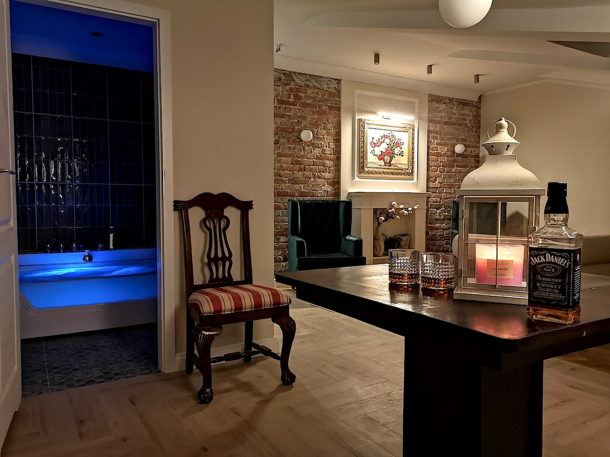 An elegant interior space features a wooden table holding a lantern and glasses. A vintage chair with striped upholstery is positioned nearby. The background reveals a well-lit seating area with a blue-tinted jacuzzi and warm-colored brick walls, creating a cozy and inviting environment.
