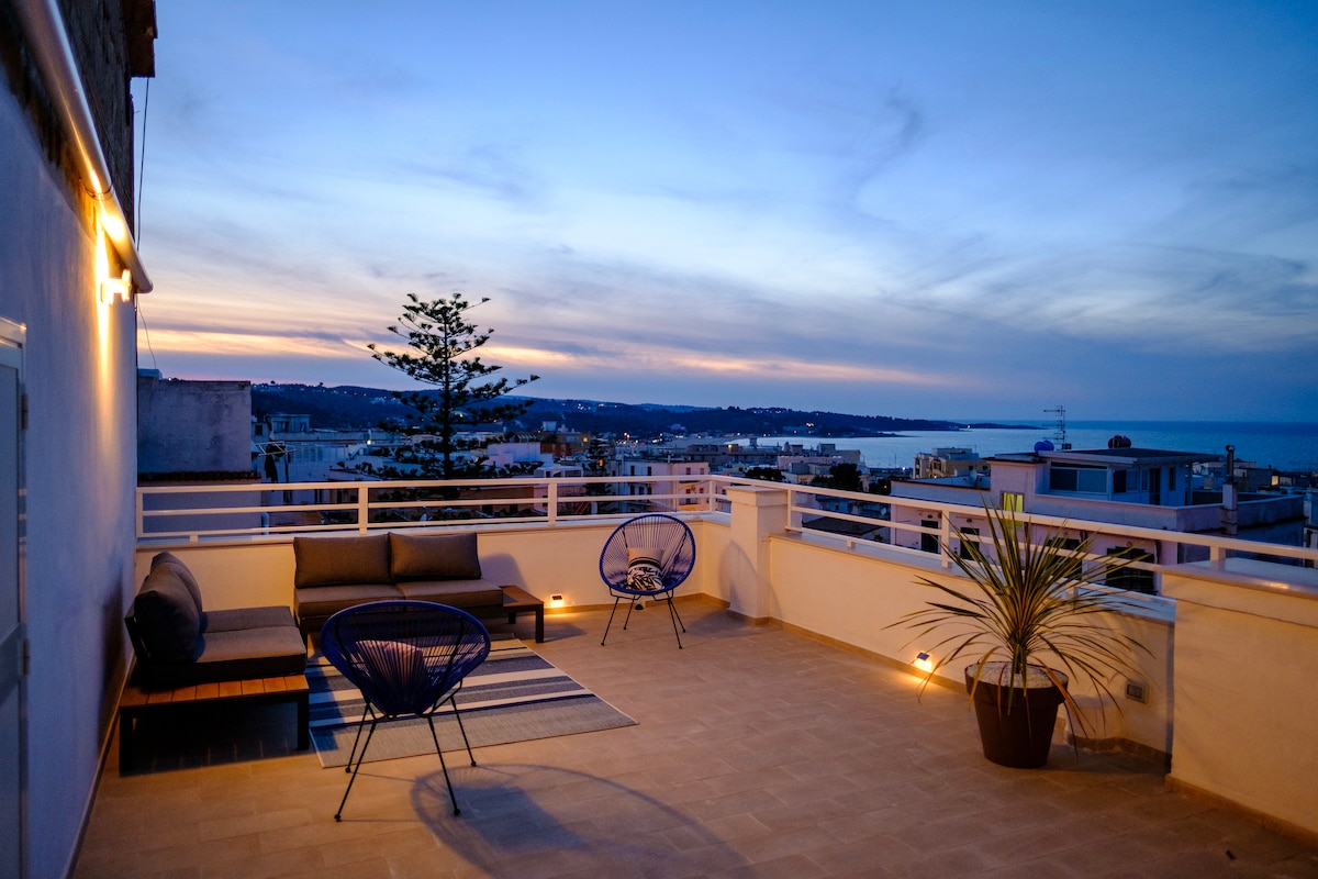 An exclusive private terrace is displayed at dusk, featuring comfortable seating and a cozy rug. Soft lighting enhances the serene atmosphere, while views of the city and sea can be seen in the background. A potted plant adds a natural touch to the space.