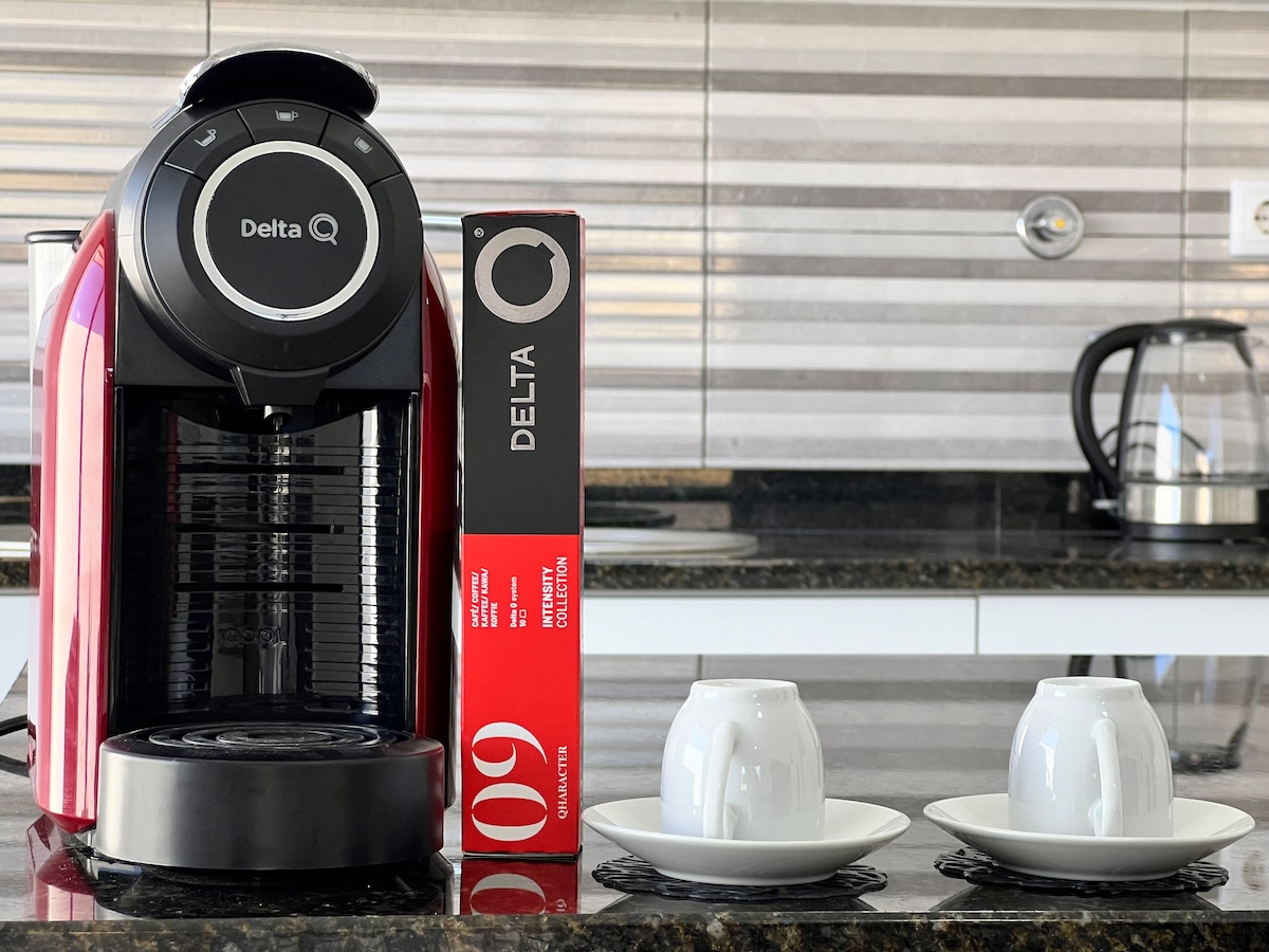 A coffee machine is displayed on a marble countertop, accompanied by two white cups on saucers. A box of Delta coffee is positioned beside the machine. A kettle is visible in the background, and the overall setting presents a modern and functional kitchen area.