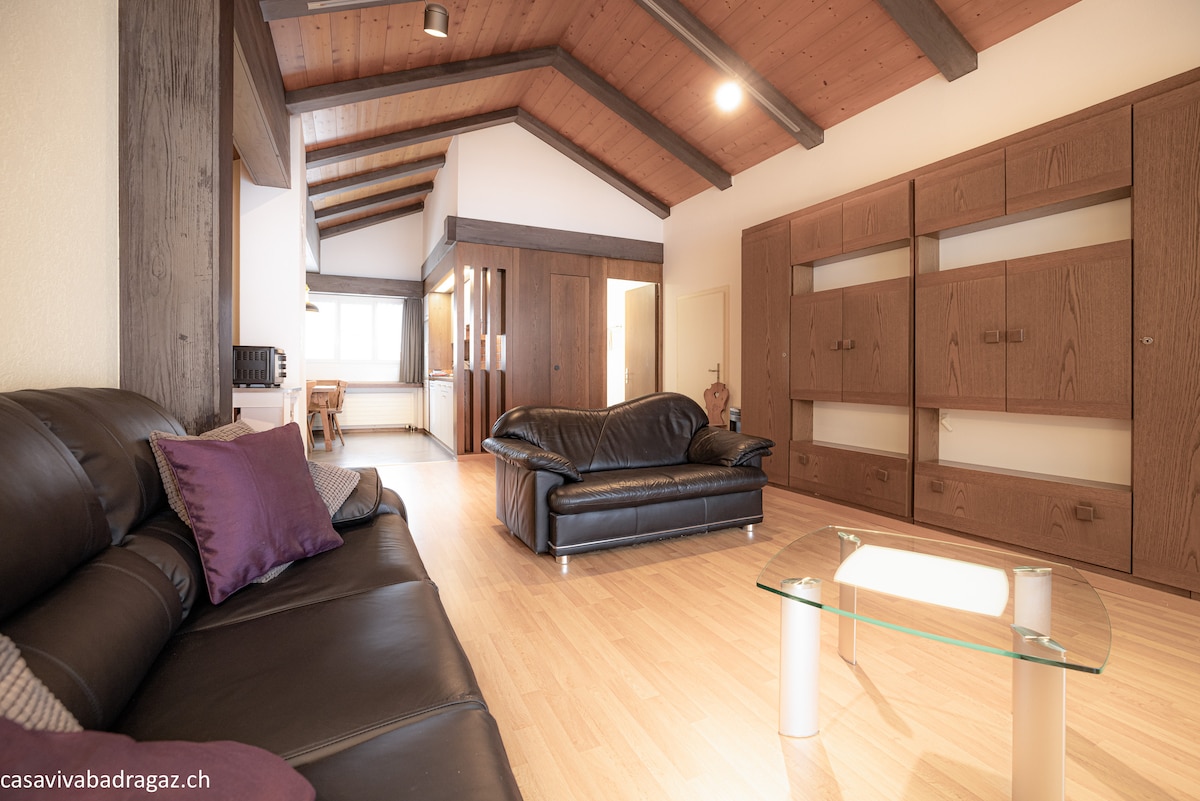 An open living area features a set of dark leather sofas with purple accent pillows, arranged around a glass coffee table. Natural light fills the space, highlighting the wooden beams and built-in cabinetry along the walls.