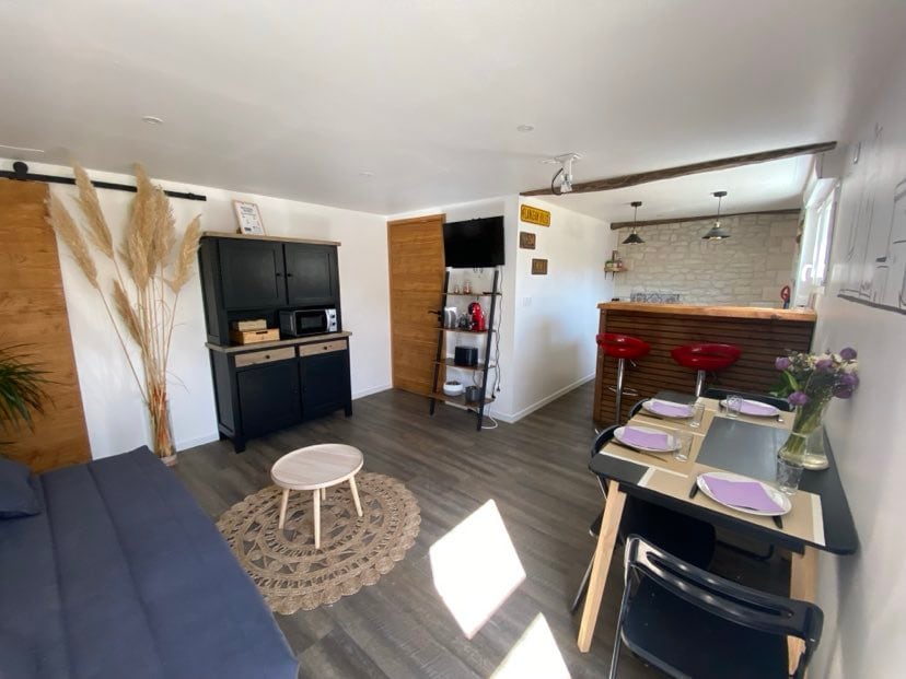 An open living area features a dark cabinet, a microwave, and a television mounted on the wall. A circular coffee table sits on a woven placemat, accompanied by a seating area with a dark sofa. A dining table is set with four chairs and a vase of flowers.