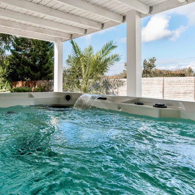 A spacious hot spring jacuzzi is situated under a covered area, featuring warm water with gentle bubbles. A palm tree is visible in the background, and natural light enters through the open space, contributing to a relaxing atmosphere.