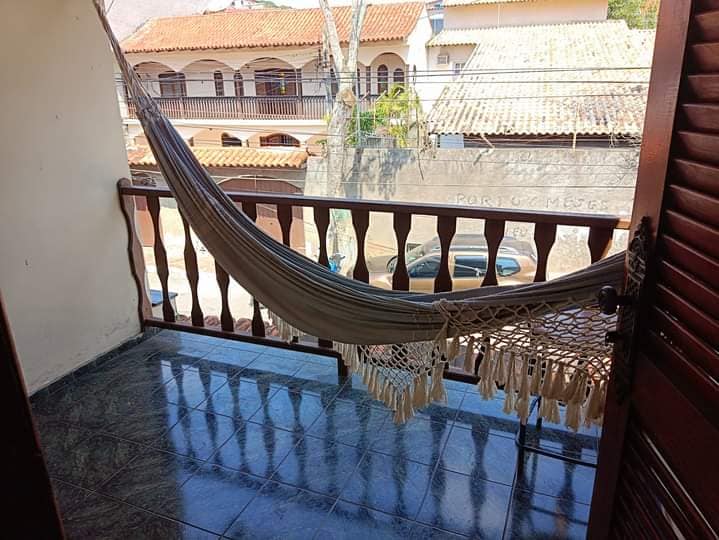 A private balcony features a cozy hammock suspended between wooden railings. The floor is tiled and reflective, adding a sense of openness. In the background, traditional architecture with terracotta roofs is visible, providing a glimpse of the surrounding neighborhood.